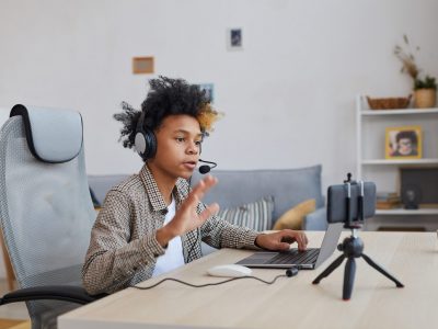 Side view portrait of teenage African-American boy wearing headset and waving at camera while streaming video games at home, young gamer or blogger concept, copy space